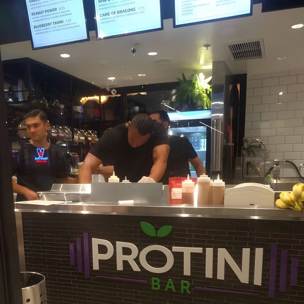 Protini Bar counter with two employees preparing acai bowls, menu screens above, and bananas on the counter.