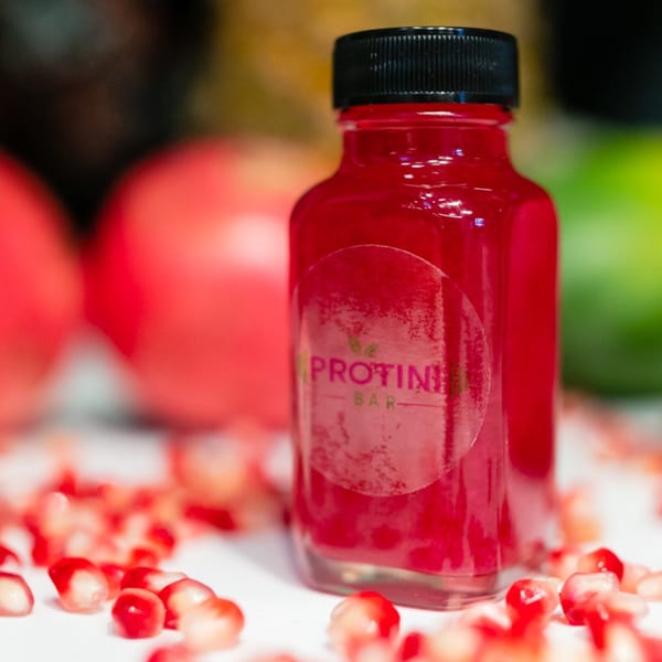 Protini Bar branded juice bottle filled with red liquid, surrounded by pomegranate seeds.