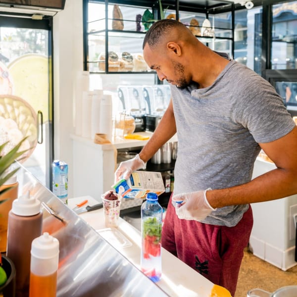 A Protini Bar employee in gloves pours milk into an acai bowl behind the counter.