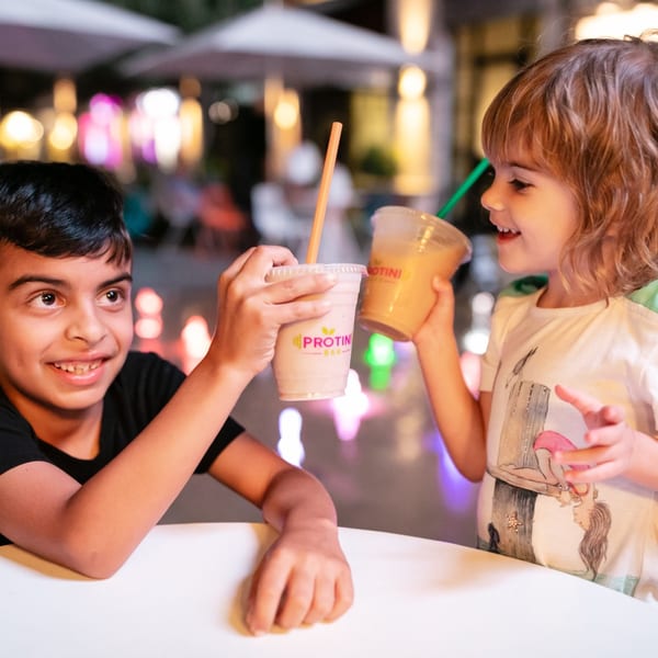 Two happy children clinking Protini Bar smoothies, one vanilla and one orange, at an outdoor table.