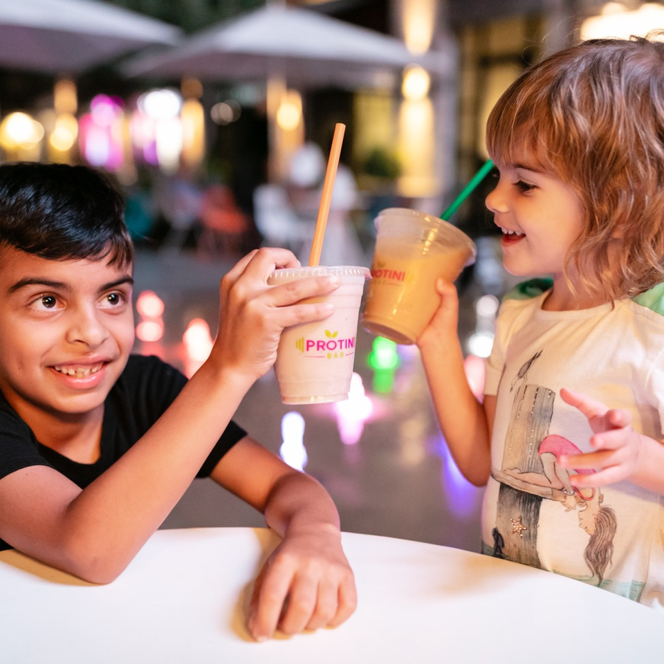 Two happy children clinking Protini Bar smoothies, one vanilla and one orange, at an outdoor table.