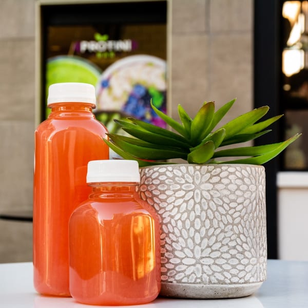 Two orange juice bottles and a potted succulent on a white table outside Protini Bar.