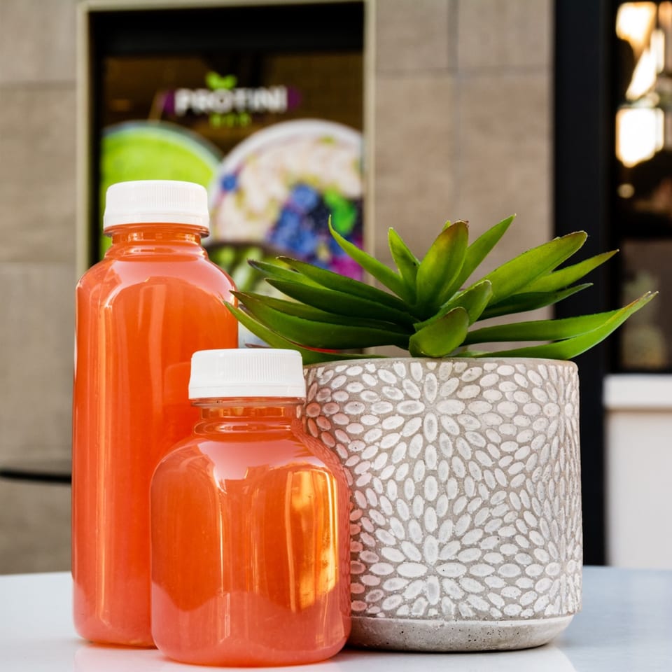 Two orange juice bottles and a potted succulent on a white table outside Protini Bar.