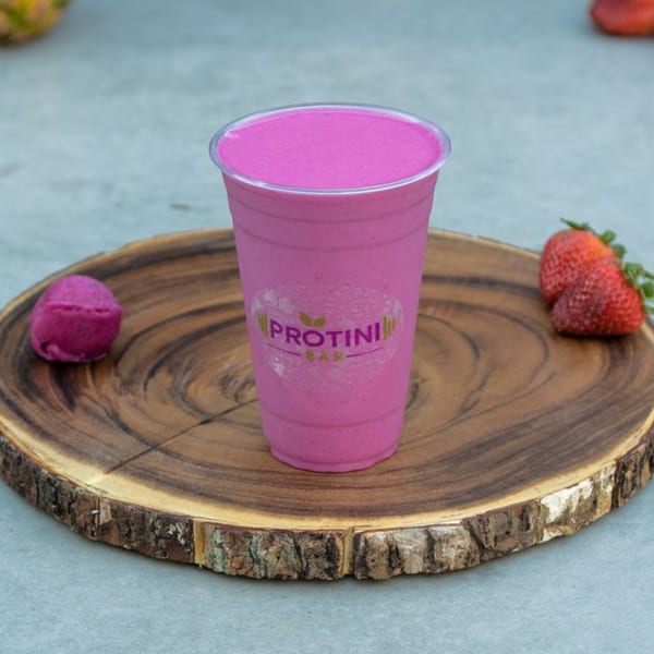 A vibrant pink Protini Bar smoothie in a clear cup, with strawberries and a berry scoop on a wooden board.
