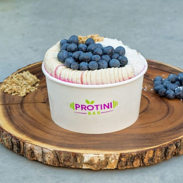 Protini Bar acai bowl with blueberries, banana slices, granola, and shredded coconut on a wooden board.