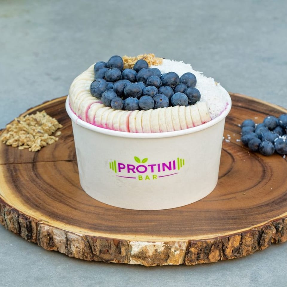 Protini Bar acai bowl with blueberries, banana slices, granola, and shredded coconut on a wooden board.