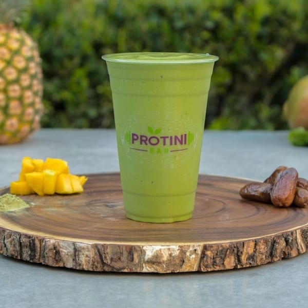 A green Protini Bar smoothie in a clear cup, surrounded by mango, dates, and pineapple.